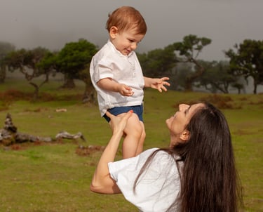 Mother lifting smiling toddler up, green grass, foggy forest and laurel trees in background, joy and play.