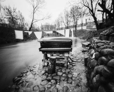 A pinhole captures a black and white photo of a piano gracefully placed on the side of a serene river