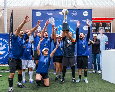 A winning amateur soccer team in blue jerseys celebrates their victory by lifting a silver trophy on a turf field.