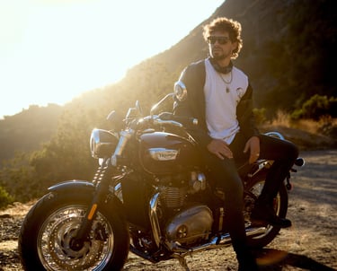 Hombre con gafas de sol y chaqueta de cuero, sentado en una motocicleta Triumph, al atardecer en un paisaje montañoso
