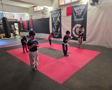 Young children practice martial arts bows on a red and black foam mat in a combat sports gym.
