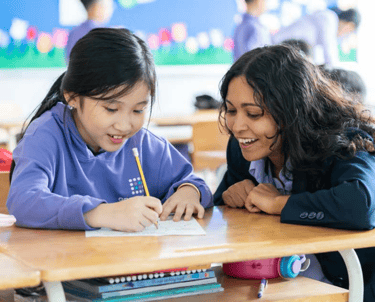 A female teacher  to the right of a student, teacher her with a smile on her face. The student is working happily, writing.