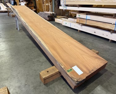 A long, single-slab live edge wood board with natural grain resting on blocks in a lumber warehouse.