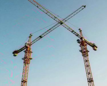 A low-angle shot of three construction cranes against a bright blue afternoon sky. The steel arms cross in a geometric pattern, symbolizing strength and progress.