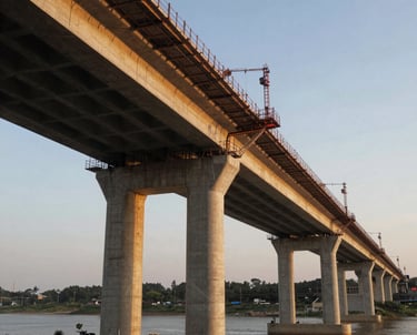 A view of a modern bridge under construction at sunset, with a golden glow hitting the concrete pillars. The composition highlights the integration of infrastructure into the natural landscape.