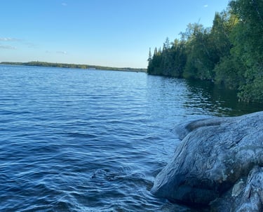 A calm lake with rocky shoreline and lush green trees under a clear sky.