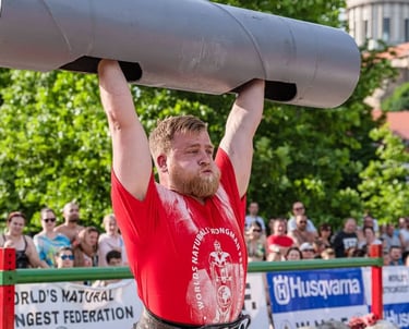 A strongman competitor in a red shirt performs a heavy log press lift at an outdoor competition.