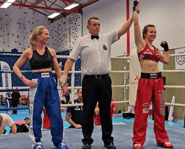 Referee raising the hand of a female kickboxer in victory inside a competitive boxing ring.