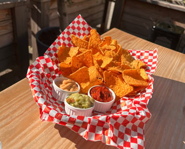 A basket of crunchy tortilla chips served with salsa, guacamole, and cheese dip on an outdoor patio table.