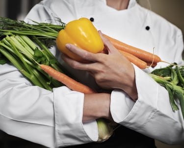 A professional chef in a white uniform holding fresh carrots and a yellow bell pepper.