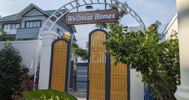 A decorative archway with a wooden sign reading 'Wellness Homes' stands above yellow gates. Lush gre