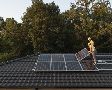 Professional technicians installing residential solar panels on a dark tiled roof near a forest.