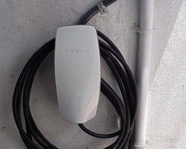 A white Tesla Wall Connector electric vehicle charger mounted on an exterior wall with a coiled black charging cable.