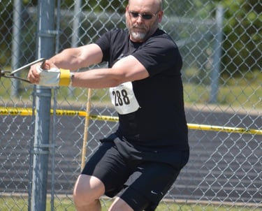 A male athlete wearing black athletic gear and sunglasses competes in a hammer throw event.