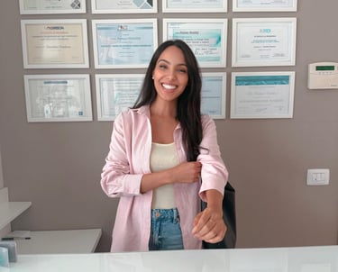 A smiling female receptionist in a pink shirt standing at a dental clinic desk with medical diplomas on the wall.