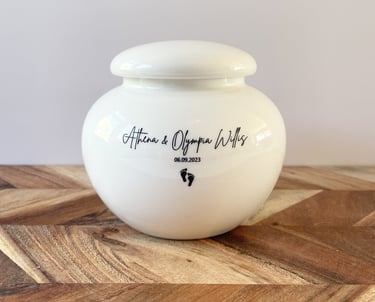 This is a rounded ivory ceramic urn engraved with the text Athena & Olympia Willis, and adorned with a small footprint icon