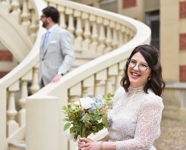 Une mariée sourit face caméra, son mari est derrière sur un escalier il descend
