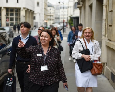 Une guide pointe du doigt un bâtiment en souriant, suivie d'un groupe de visiteurs. Ils marchent dans la rue