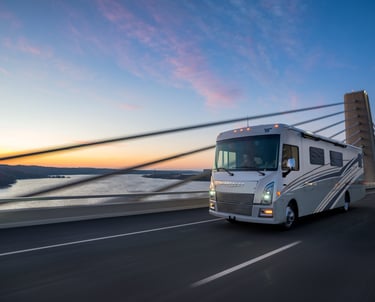 A Winnebago Class A motorhome traveling across a suspension bridge over water during a sunset.