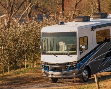 A white Fleetwood Bounder Class A motorhome driving through a scenic rural apple orchard at sunset.