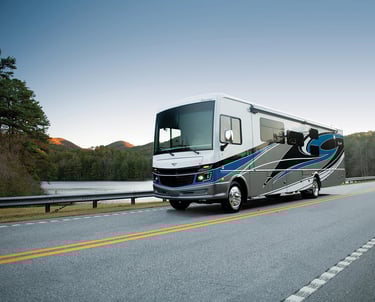 A luxury Class A motorhome driving along a scenic mountain road beside a peaceful lake at sunset.