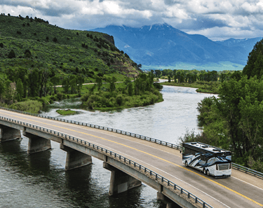 A luxury Class A motorhome drives across a bridge over a scenic river with mountain views.