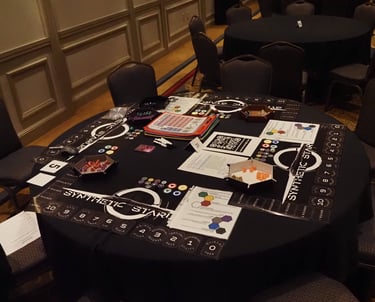 A table set up to run a game of Synthetic Starlight at Gen Con 2025. Dice, dice trays, and playmats.