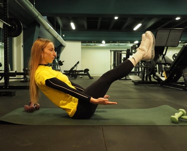 Una donna in maglia gialla esegue l'esercizio 'boat pose' su un tappetino da yoga in palestra, concentrandosi sull'equilibrio