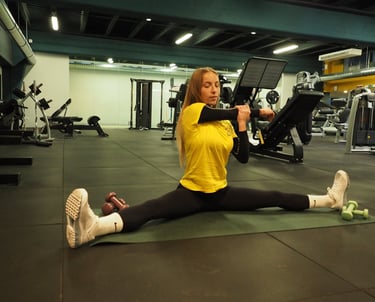 Una donna in maglia gialla esegue uno stretching in spaccata seduta, allungando il braccio per un esercizio di mobilità,