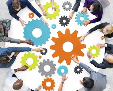 A group of people seated around a table are collaboratively arranging colorful gear-shaped pieces, symbolizing teamwork