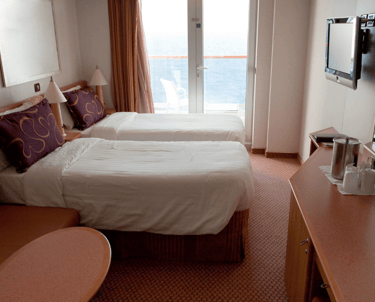 Cruise-ship cabin with two twin beds, a balcony door overlooking the ocean, wall-mounted TV, small sofa, and a desk