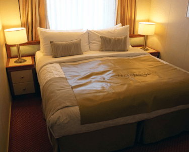 Cozy cruise-ship stateroom with a king bed, two bedside tables and lamps, and a curtained window above the headboard