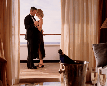 Formal couple dancing on a cruise-suite balcony with the ocean beyond, seen through curtains, with champagne and flutes