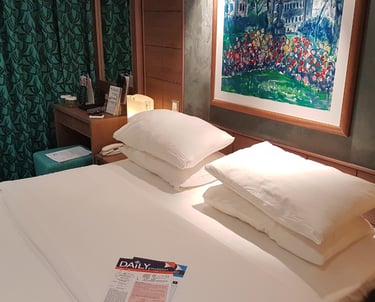 Cozy cruise-ship stateroom with a made king bed and stacked pillows, bedside lamps, green patterned curtains, a framed painti