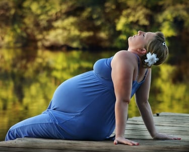 A pregnant woman in a blue dress sits on a wooden dock by a peaceful lake at sunset.