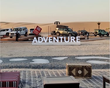 An outdoor car show setup in the desert featuring SUVs, a stage, and a large "ADVENTURE" sign.