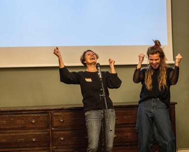 Two women cheering behind a microphone, one public speaking trainer and one participant.