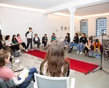 Woman walking down a red carpet toward a microphone, surrounded by seated women at a speaking event.