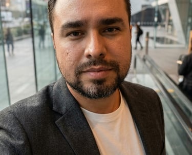 Professional man in a gray blazer and white t-shirt standing in a modern office building with city views.