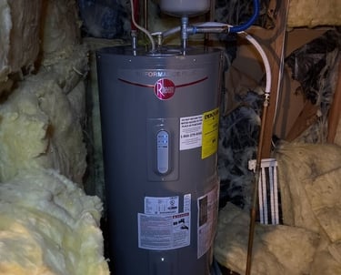 A Rheem Performance Plus electric water heater installed in an attic surrounded by fiberglass insulation.