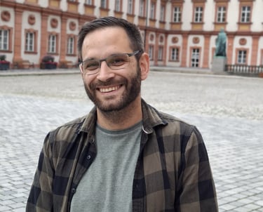 Inhaber, Patrick Angerer der freundlich in die Kamera guckt und in Bayreuth steht im Hintergrund altes Schloss in Bayreuth