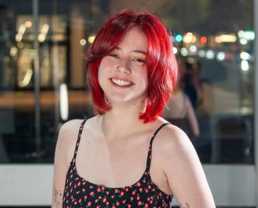 Smiling young woman with vibrant red bob haircut wearing a floral sundress in a city setting.