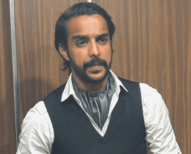 A man with a mustache wearing a black vest, cravat, and white shirt standing against a wood wall.