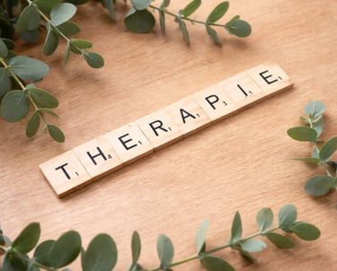 Wooden letter tiles spelling Therapie surrounded by green eucalyptus leaves on a desk.