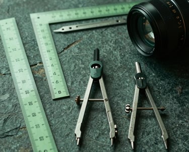 A collection of architectural drawing tools—rulers, compasses—arranged neatly on a dark stone surface. Forest green tones.