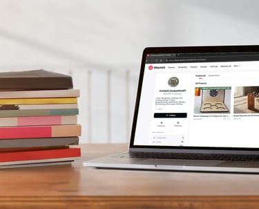 A stack of hardcover books sits next to an open laptop on a wooden desk showing a design website.