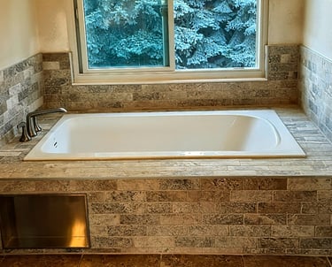 A modern white soaking tub surrounded by grey stone tile and a window overlooking snow-covered pine trees.