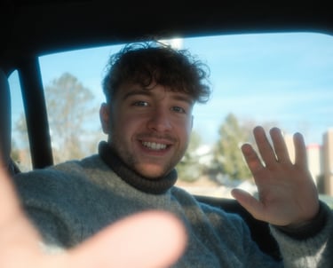 Smiling young man with curly hair waving while sitting in the back seat of a car.