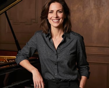 A professional portrait of a smiling woman in a dark charcoal shirt, standing next to a grand piano in a studio with rich bronze lighting.