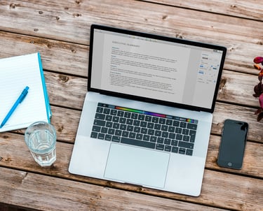 Un portátil que muestra un documento profeison, con un cuaderno, un celular y un vaso de agua.